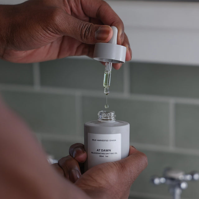 Close-up of the glass dropper extracting the golden At Dawn Rejuvenating Daytime Oil from the bottle in a bathroom setting.