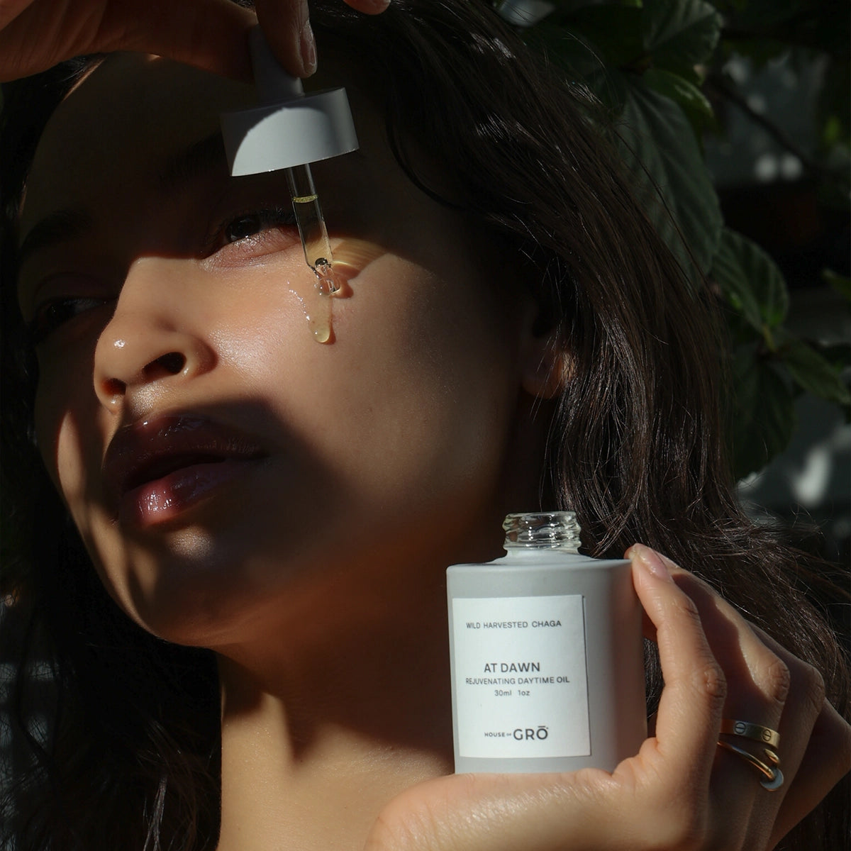 Woman applying a drop of House of GRŌ At Dawn Rejuvenating Daytime Oil directly to her cheek, showing a glowing finish in the sunlight.